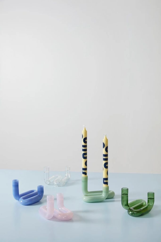 Minimalist still‑life: pastel glass sculptures and two tall blue‑patterned candles on a pale blue surface.