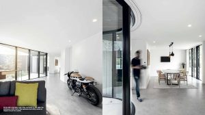Open-plan modern home interior with a tan motorcycle by a curved glass wall; dining area on the right and a lounge area on the left. A blurred person walks toward the dining space.
