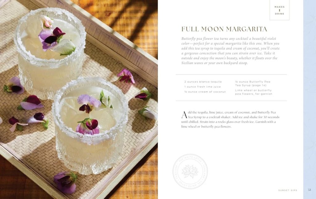 Two margarita-style cocktails with sugar-rimmed glasses and flower petals on a wooden tray, sunlight casting warm shadows.