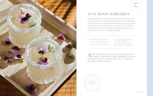 Two margarita-style cocktails with sugar-rimmed glasses and flower petals on a wooden tray, sunlight casting warm shadows.