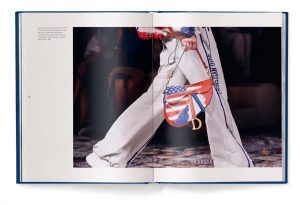 Open fashion book showing a model's white pants and a red, white, and blue handbag with a Dior charm on a runway scene.