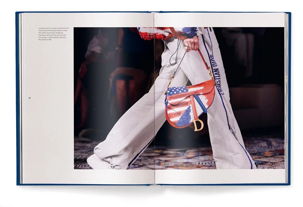 Open fashion book showing a model's white pants and a red, white, and blue handbag with a Dior charm on a runway scene.