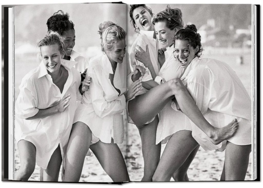 Group of women on a beach, wearing oversized white shirts, laughing and lifting their legs in a playful pose.