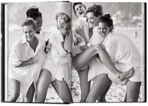 Group of women on a beach, wearing oversized white shirts, laughing and lifting their legs in a playful pose.