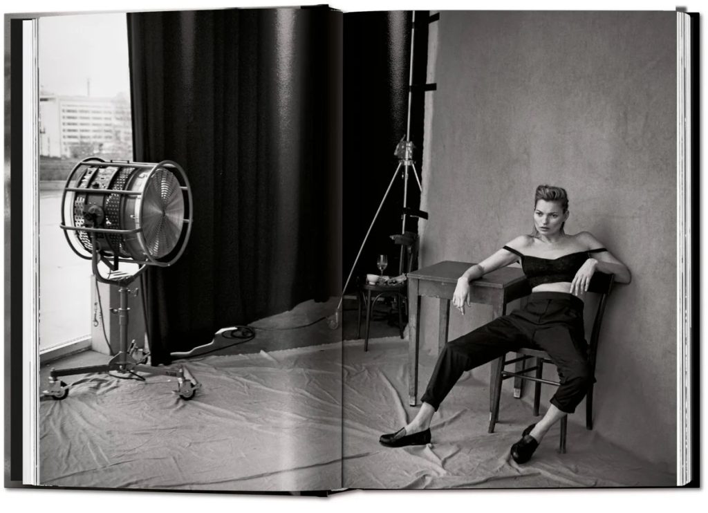 Model posing in a studio with an off-shoulder top, black pants, and loafers while seated at a table; vintage photography backdrop and large studio light to the left.