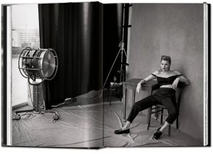 Model posing in a studio with an off-shoulder top, black pants, and loafers while seated at a table; vintage photography backdrop and large studio light to the left.