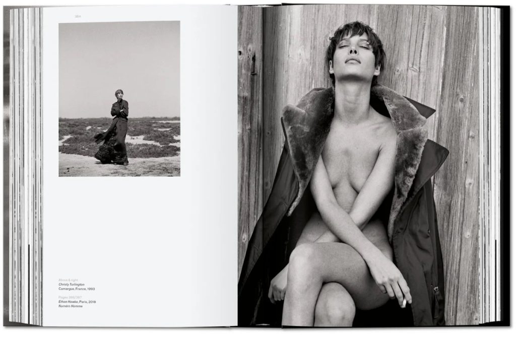 Open magazine spread with a black-and-white fashion photo: a topless model sits against a wooden wall wearing a fur-cly coat, on the right page; left page shows a figure on a beach.
