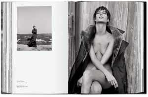 Open magazine spread with a black-and-white fashion photo: a topless model sits against a wooden wall wearing a fur-cly coat, on the right page; left page shows a figure on a beach.